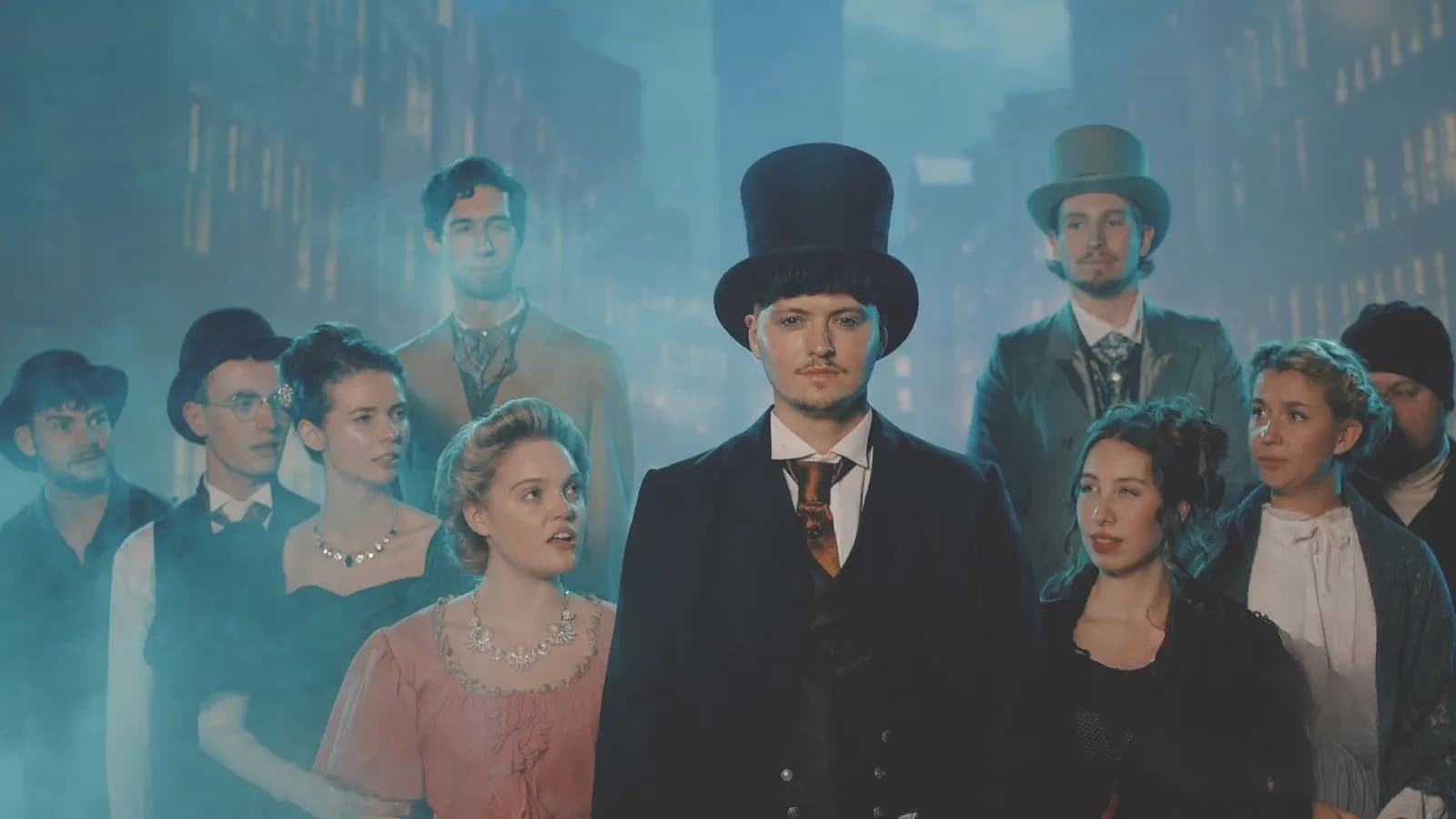 A group of actors in historical costumes, with men in top hats and suits, and women in period dresses, appearing in a misty, blue-lit setting.